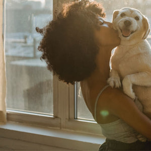 Black woman standing in front of a window holding and kissing a small beige dog who is smiling